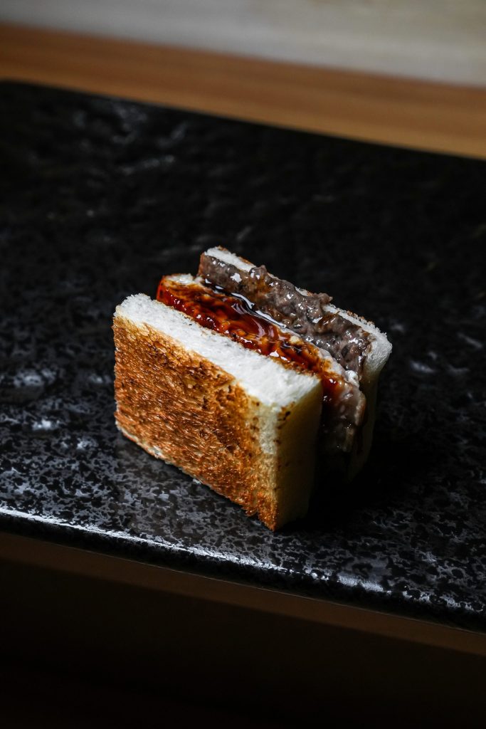 Wagyu sando on toasted white bread with a glossy sauce, on a black stone plate